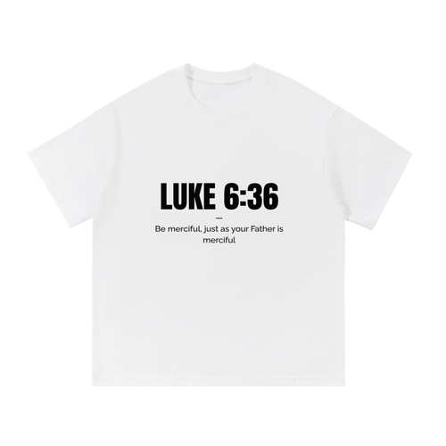 A white short-sleeve crewneck t-shirt with black printed text on the chest reading 'LUKE 6:36' and 'Be merciful, just as your Father is merciful'.